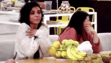 two women are sitting at a table with a bowl of bananas and fruit .