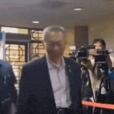 a man in a suit and tie is walking through a room with cameras and a red ribbon .