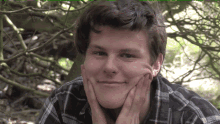 a young man in a plaid shirt is smiling with his hands on his chin