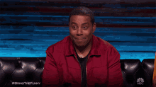 a man in a red jacket is sitting on a black couch with a nbc logo in the corner