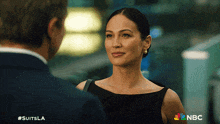 a woman in a black dress is looking at a man in a suit and the nbc logo is visible