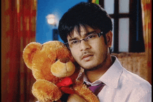 a man wearing glasses is holding a teddy bear with a red heart in its mouth
