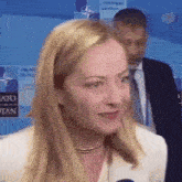 a woman in a white jacket is talking to a microphone while a man in a suit stands behind her .