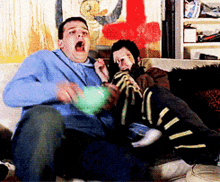 two men are sitting on a couch playing a video game with their mouths open