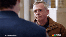 a man in a brown jacket is talking to another man in a blue shirt with the nbc logo on the bottom right