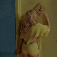 a woman in a yellow shirt and shorts stands in front of a door with a sign that says 2 on it
