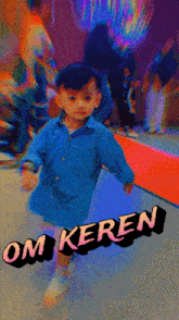 a little boy in a blue shirt is standing in front of a sign that says " om keren "