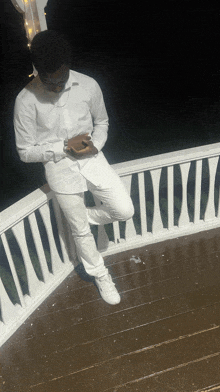a man in a white shirt and white pants sits on a balcony looking at his phone
