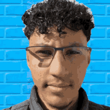 a man with curly hair wearing glasses and a black shirt stands in front of a blue brick wall