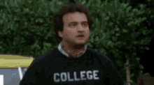 a man wearing a black t-shirt with the word college on it is standing in front of a bush .