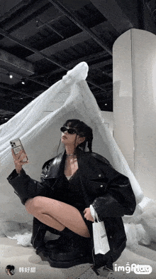 a woman in a black leather jacket is kneeling down holding a phone and a bag