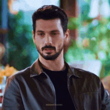 a man with a beard wearing a leather jacket and a black shirt is looking at the camera .