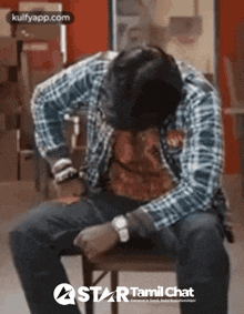 a man in a plaid shirt sits on a chair with his head on his knees