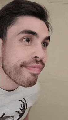 a man with a beard is wearing a white shirt with a deer on it and making a funny face .