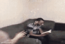 a man is sitting on a couch reading a book while another person holds a remote control .