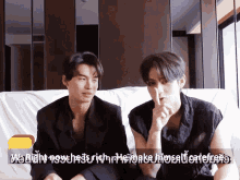 two men are sitting on a couch and one of them has his finger on his mouth