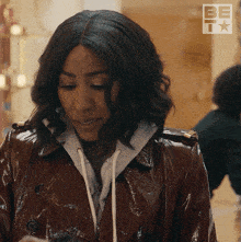 a woman wearing a brown trench coat and a grey hoodie is standing in front of a be tv sign