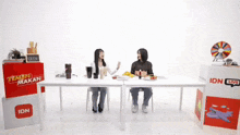 two people sit at a table with a sign that says idn live on it