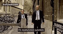 two men in suits and ties are walking down a street with bicycles parked on the sidewalk .