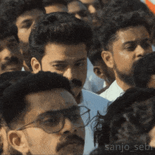 a man with glasses and a mustache stands in a crowd with the name sanjo_sebi on the bottom left