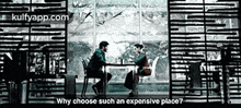 two men are sitting at a table in front of a window and talking .