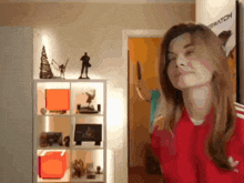 a woman in a red adidas shirt stands in front of a shelf with a poster that says overwatch
