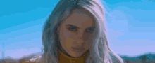 billie eilish is wearing a yellow turtleneck and looking at the camera while standing in front of a blue sky .