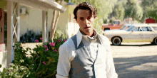 a man in a white shirt and gray vest stands in front of a yellow car
