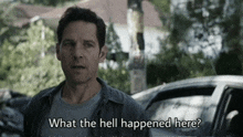 a man in a blue shirt is standing in front of a car and asking what the hell happened here .