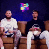 two men are sitting on a couch playing video games . one of the men is wearing a spurs shirt .