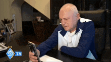 a man sits at a desk looking at a phone with a tv logo in the corner