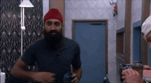 a man with a beard and a red turban is standing in a room holding a cup .
