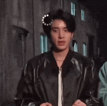 a young man wearing a black leather jacket is standing in front of a building and looking at the camera .