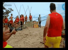 a man in a red shirt and yellow shorts stands in front of a group of people on the beach