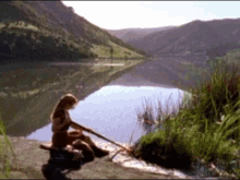 a woman sits on the shore of a lake holding a spear