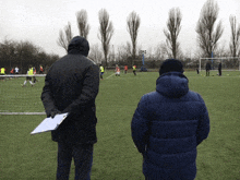 two men are watching a soccer game on a field with trees in the background