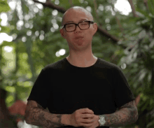 a man with glasses and a tattoo on his arm stands in front of trees