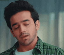 a man wearing a green plaid shirt looks down with his eyes closed