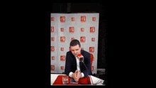 a man in a suit sits at a table with a microphone in front of a wall with ffi written on it