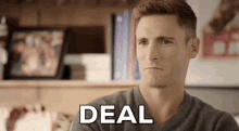 a man is sitting at a table with a bookshelf in the background and says `` deal '' .