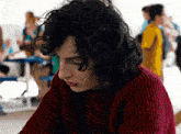 a young man with curly hair wearing a red sweater looks down