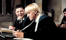 two boys are sitting at a table looking at a book and smiling