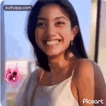 a woman in a white tank top is smiling and looking at the camera with a pink flower in the background .