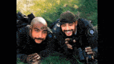 two men are laying in the grass with one holding binoculars and the other a compass