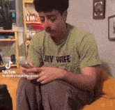 a man in a green shirt that says my wife is sitting on a couch looking at his phone .