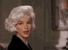a woman with blonde hair and a black jacket is looking at the camera in a room .