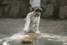 a woman in a white robe is kneeling on a rock near a pond