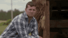 a man in a plaid shirt is standing in front of a wooden shed with corn on the cob .
