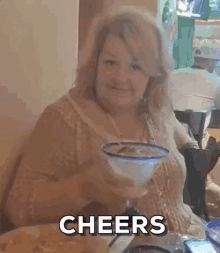 a woman is sitting at a table with a margarita in her hand and the words cheers behind her .