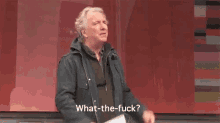 a man in a jacket is standing in front of a red wall and saying `` what the fuck ? ''